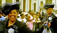 Two women wearing their Sunday best move to the music as they engage in the movement of the second line down a crowded street.