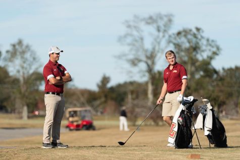 Golf coach Cory Amory stands looking at athlete holding a golf club and golf bag on the green