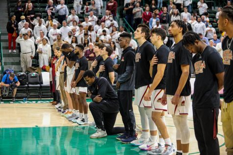 mens basketball team stands for national anthem on court while one athlete bends the knee