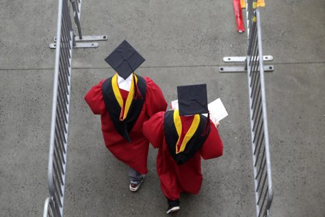 Two graduating students, wearing graduation cap and gowns, walk down pavement.