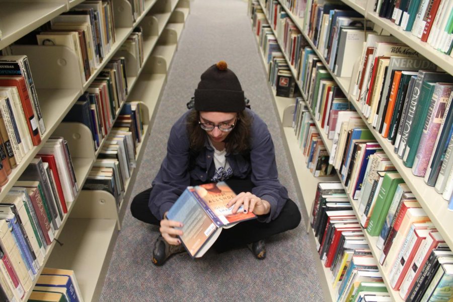 A photo of someone sitting on the floor of a library reading the back of a book.