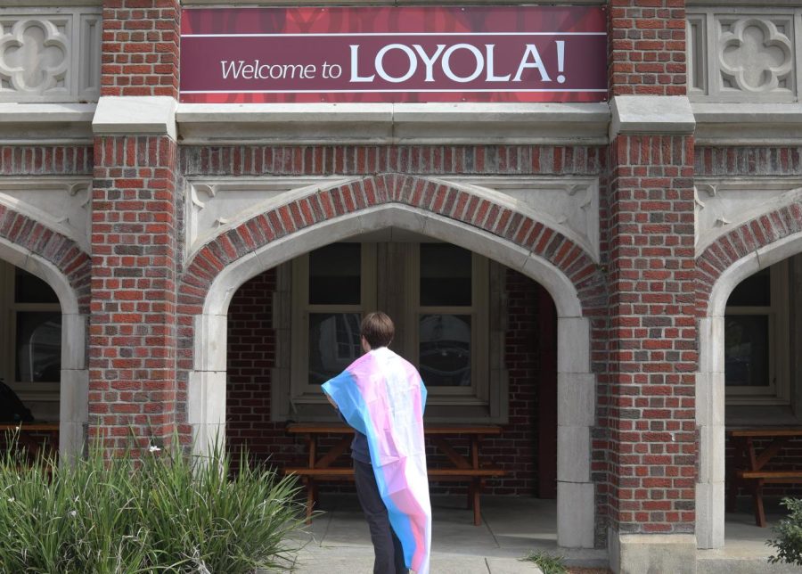 A picture of someone with a trans flag draped over them standing under a sign that says welcome to Loyola.